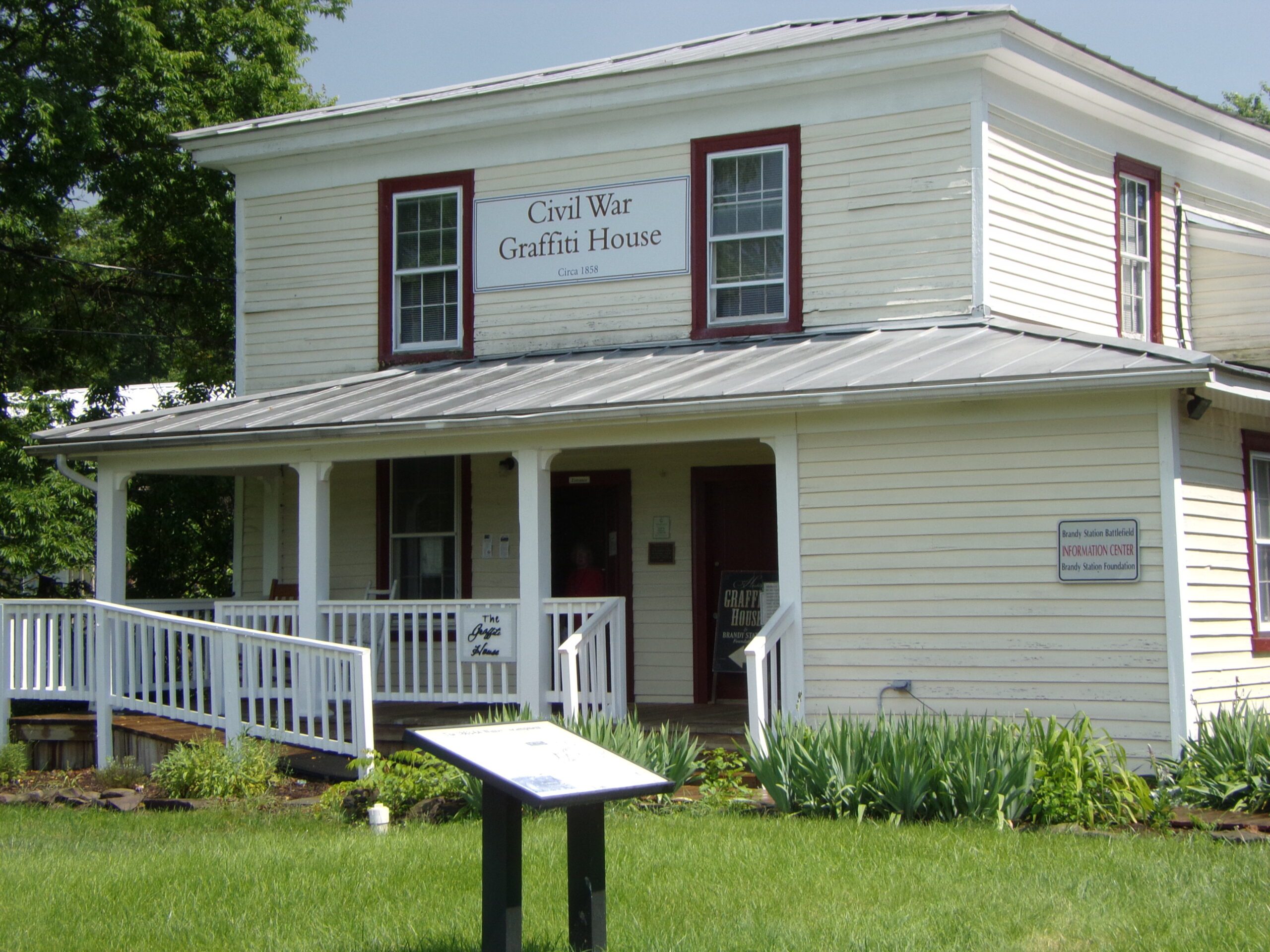 A white, two-story wooden house with burgundy trim, featuring a sign that reads "Civil War Graffiti House, circa 1858." The building has a porch with a ramp and steps, a historical marker in the yard, and some green shrubs and plants in front.