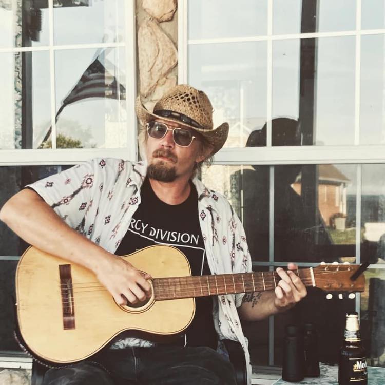 A man wearing a straw hat, sunglasses, and a patterned shirt over a t-shirt sits in front of a window, strumming an acoustic guitar. A can and binoculars are visible on the adjacent surface. An American flag is reflected in the window.
