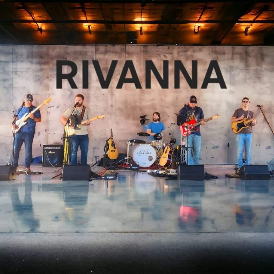 A five-member band performs on an indoor stage with instruments including guitars, drums, and a keyboard. The wall behind them has a large "RIVANNA" sign. Various lighting fixtures are mounted on the ceiling above the stage.