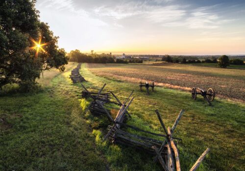 A scenic countryside landscape at sunrise, featuring a split-rail fence leading into the distance across grassy fields. Historical cannons are positioned on either side of the fence line, with a clear sky and rising sun casting a warm glow over the scene. Image