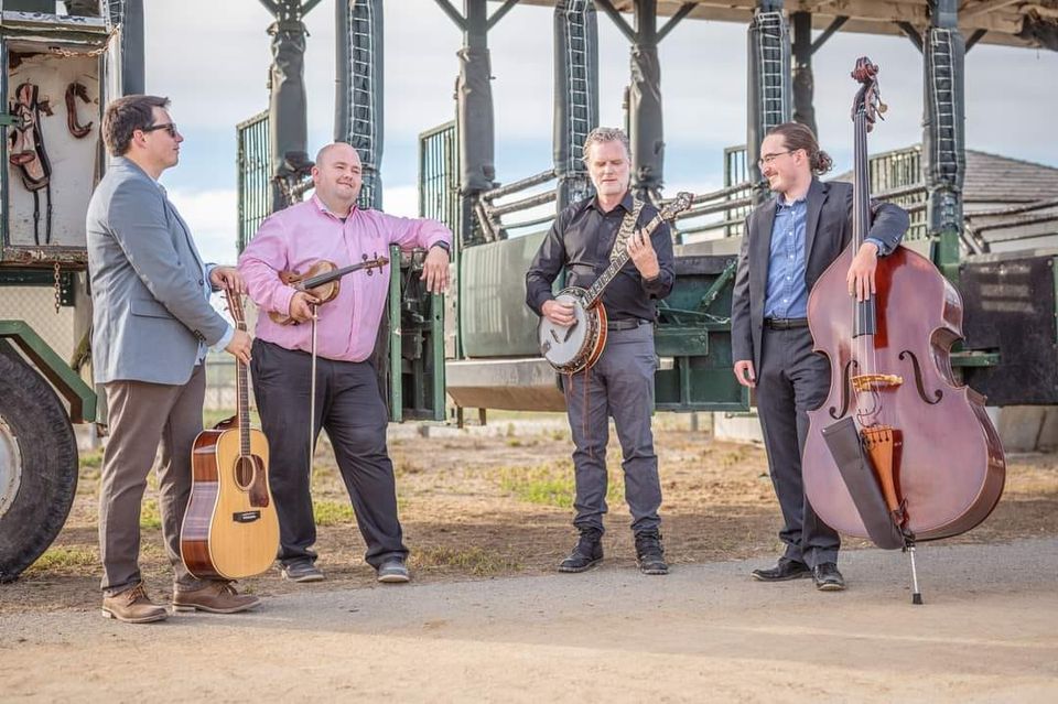 Four men are standing outside near a fenced area, each holding a musical instrument. From left to right, they have a guitar, a violin, a banjo, and an upright bass. They appear to be engaged in conversation and ready to perform.