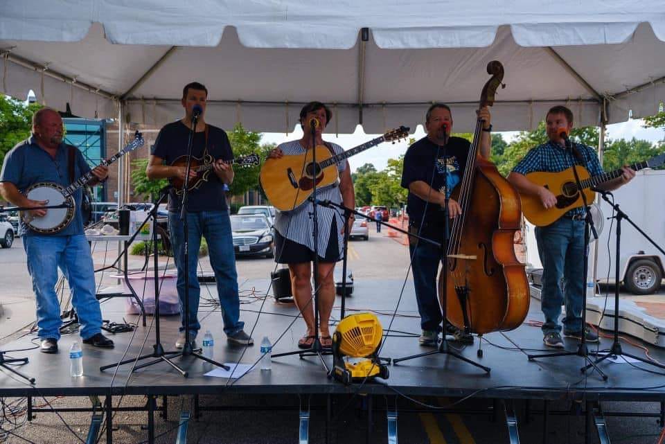 A five-member band performs under a white tent. The musicians are playing various instruments, including a banjo, mandolin, guitar, double bass, and an additional guitar. They stand on a stage with microphones and a few water bottles nearby. Trees and cars are visible in the background.