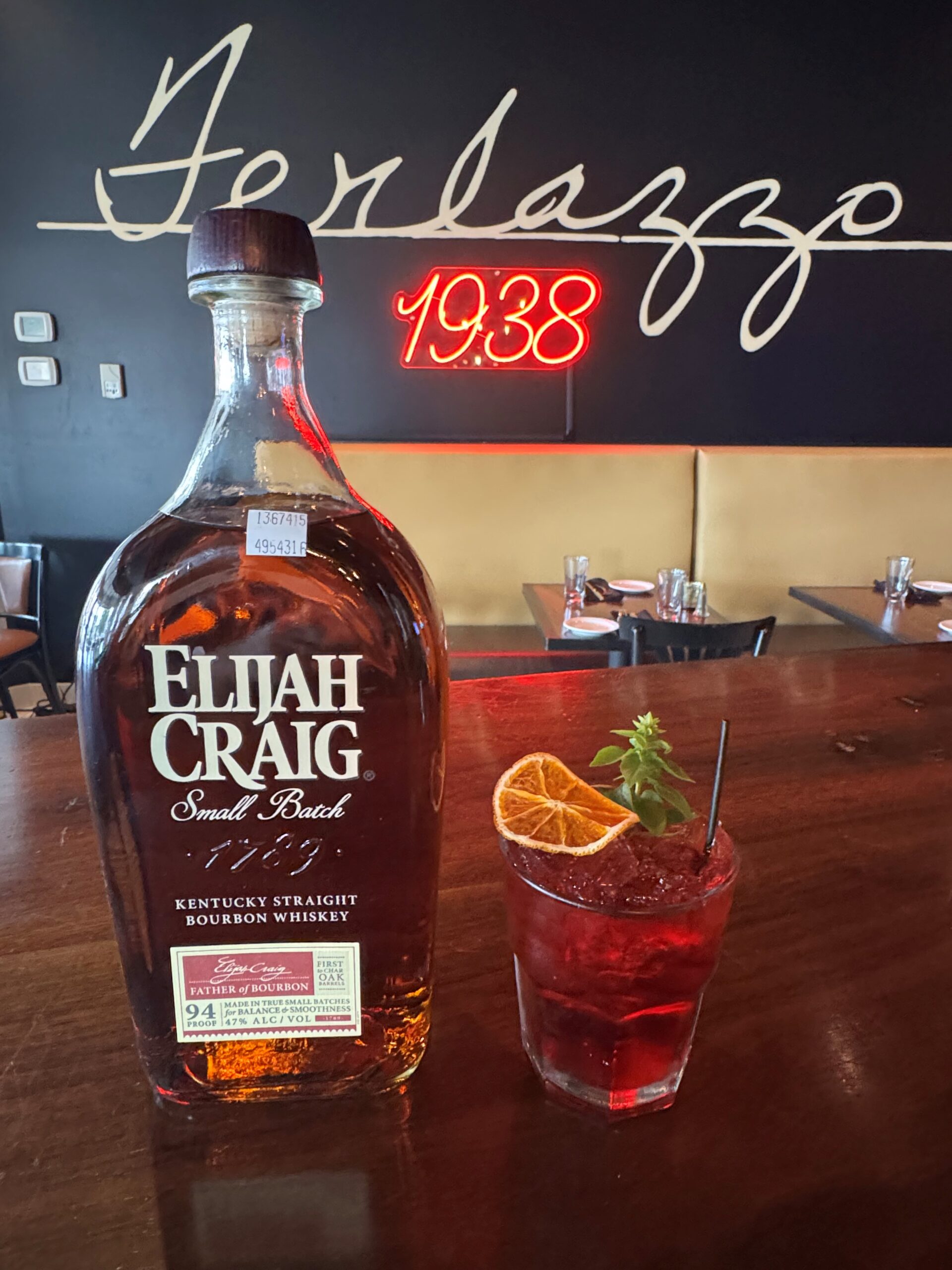 pink cocktail garnished with a dried lemon and basil leaves and a bottle of Elijah Craig next to it