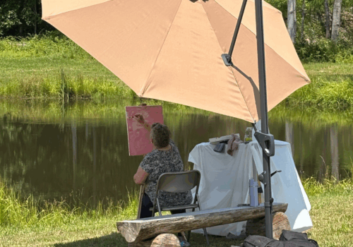 woman painting under an umbrella in front of a lake Image