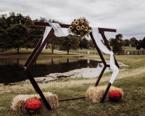 A hexagonal wedding arch stands by a pond, decorated with sunflowers, white draped fabric, and surrounded by bundles of hay. Red potted flowers are placed on the ground beside the hay. Trees and cloudy sky are seen in the background.