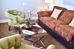 A cozy living room with a brown sofa adorned with patterned cushions, two green armchairs, and a glass coffee table. The coffee table holds a decorative tray with a vase containing white flowers and a small stack of coasters. A floor lamp stands in the corner.